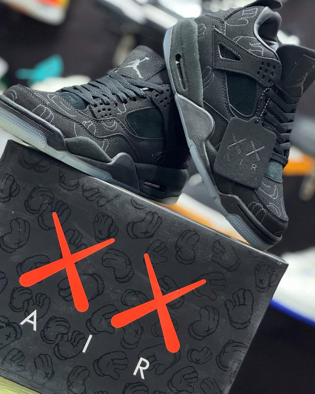 A pair of black Air Jordan 4 Retro 'XX' sneakers with gray overlays and white Jumpman logos, displayed on top of a black shoebox featuring the signature red 'XX' logo and handprint pattern.